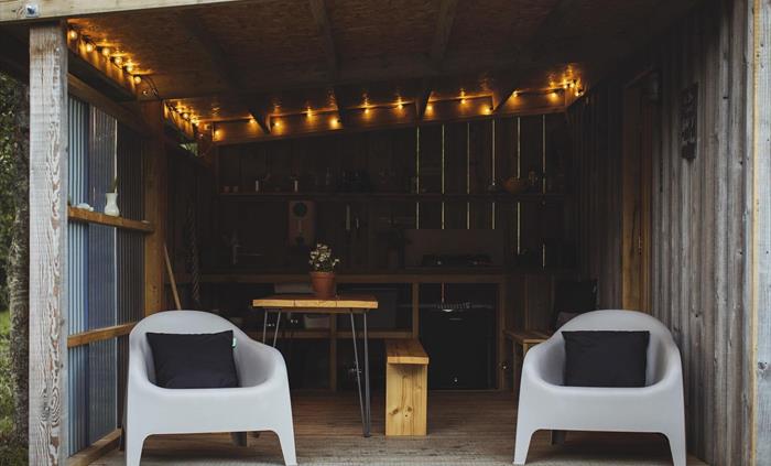A covered camp kitchen with festoon lighting above. A dining table and two lounging chairs in the foreground.